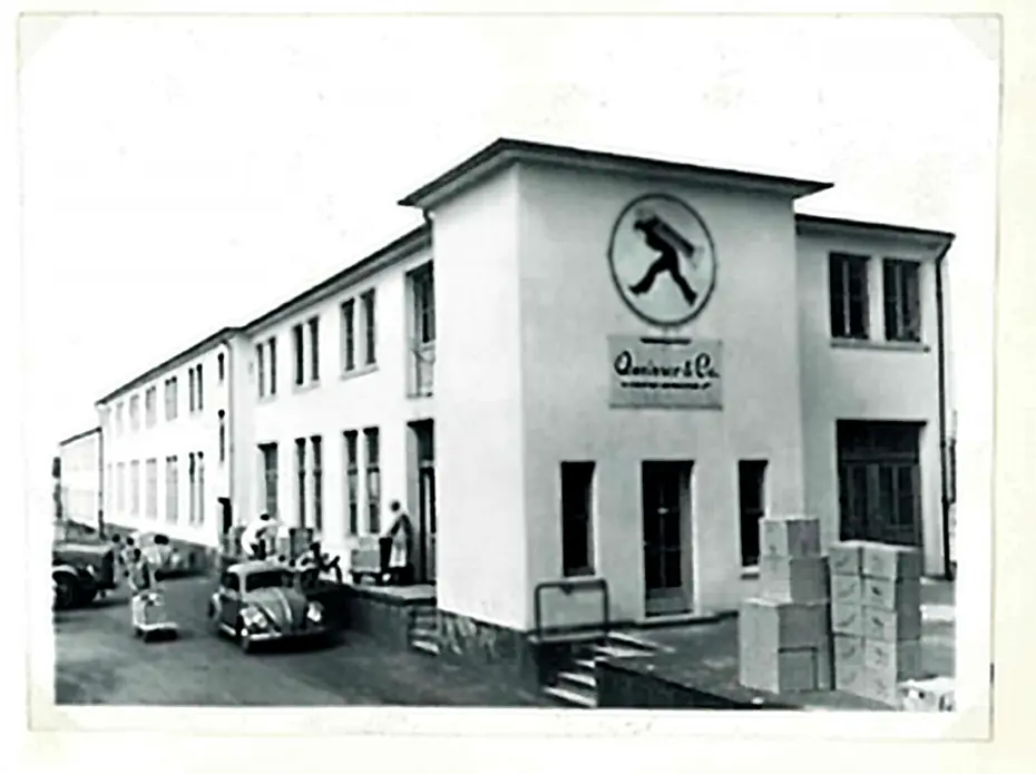 Qora va oq rangda eski binoni ko'rsatadigan foto, old tomonda logo va "Quinver & Co." yozuvi bor. O'rtada bir necha avtomobillar, tashqarida esa odamlar va qutichalar. Binoning ko'chalari va derazalarining ko'rinishi eski zamonga oid.