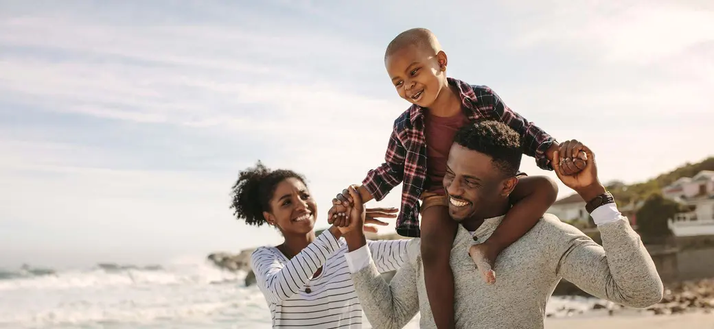 A joyful family day at the beach: a father carries his smiling son on his shoulders, while the mother holds their hands. They share laughter and happiness against a backdrop of ocean waves and a clear sky, creating a warm, loving atmosphere.