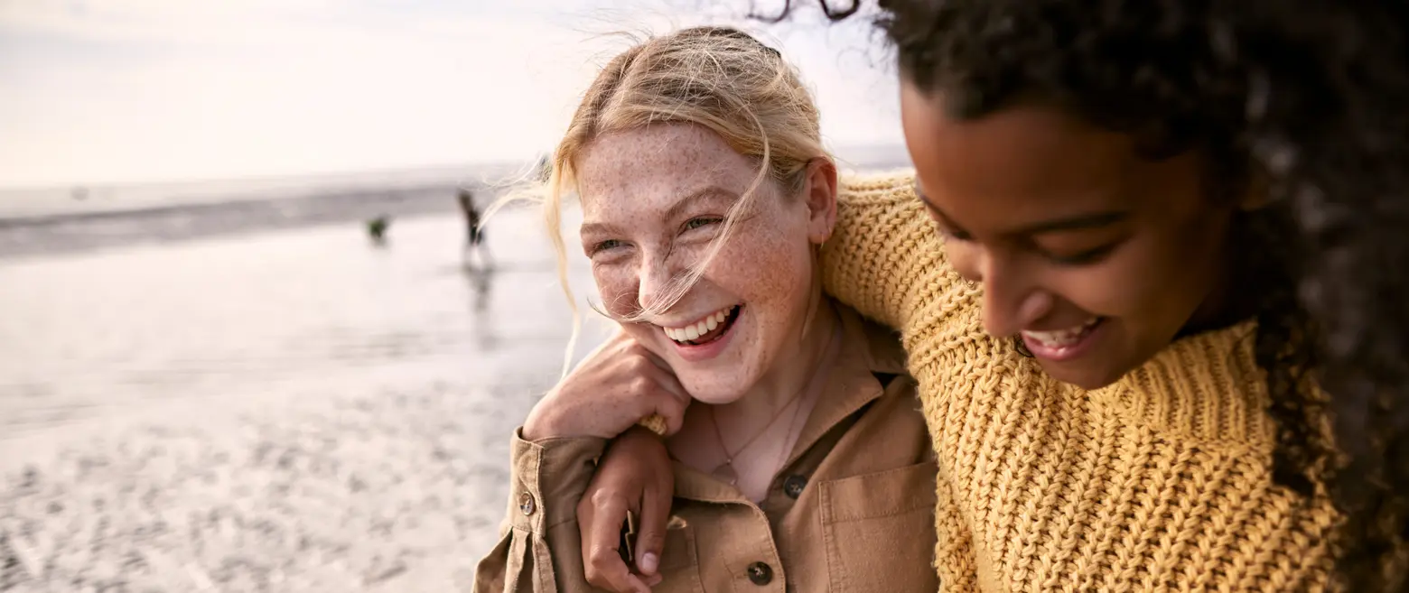 Two friends smile and share a joyful moment on a beach. One has curly hair and wears a yellow knitted sweater, while the other has straight blonde hair and freckles, dressed in a light brown top. They are close together, enjoying each other’s company amidst a serene backdrop.